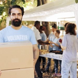 Compassionate man volunteering at a local food drive, offering non-perishable rations to the needy and homeless people. Caucasian male charity worker looking at camera while carrying donation boxes